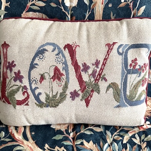 May include: A rectangular throw cushion with a cream-coloured woven fabric base. The word LOVE is embroidered in large, decorative letters. Each letter is filled with intricate floral patterns in shades of red, blue, green, and purple.