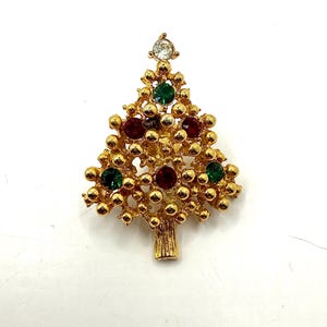 Może przedstawiać: Złota broszka w kształcie choinki z czerwonymi, zielonymi i przezroczystymi kryształowymi akcentami. Broszka ma teksturowany wzór z podstawą w kształcie pnia drzewa. Świąteczny dodatek na sezon świąteczny.