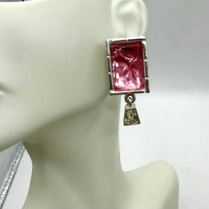 May include: A silver-toned earring with a rectangular, pink, faceted stone. The stone is set in a silver frame and has a smaller, green-toned, trapezoid-shaped dangle. The earring is displayed on a mannequin.