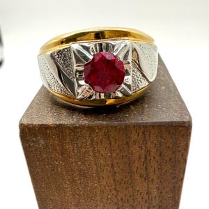 May include: A gold and silver ring with a large, round, deep red gemstone at the center. The ring has a thick band with a textured design on the sides. The gemstone is set in a square, detailed setting.