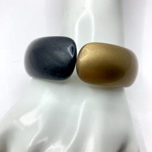 May include: Two chunky, rounded bracelets. One is a matte black, the other a metallic gold. The bracelets are displayed on a white surface. The bracelets are a bold statement piece.