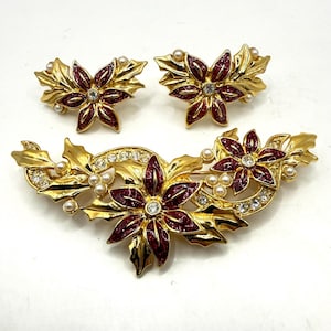 May include: A gold-toned jewelry set featuring a brooch and matching earrings. The set has a floral design with red enamel petals, clear rhinestones, and faux pearls. The brooch is approximately 3 inches long.