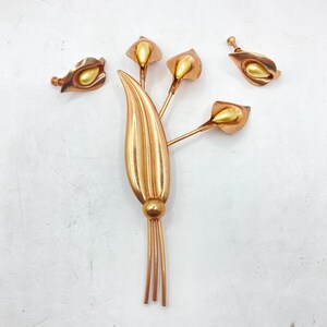 May include: A vintage copper-toned floral brooch and matching earrings set. The brooch features a stylized flower bouquet design with a large leaf and multiple calla lily-like blooms. The earrings have a similar design. The set has a gold-colored center.