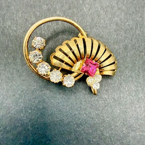 May include: A gold-toned brooch featuring a fan design with a central pink gemstone and clear, round accent stones. The brooch has a circular element with additional clear stones. The fan has vertical lines.