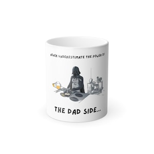 Könnte beinhalten: Weiße Keramiktasse mit einer humorvollen Illustration von Darth Vader beim Kochen. Die Tasse trägt den Text "UNTERSCHÄTZE NIEMALS DIE MACHT DER PAPA-SEITE..." in schwarzer Schrift. Das Design beinhaltet eine Karikatur von Darth Vader mit einer Schürze.