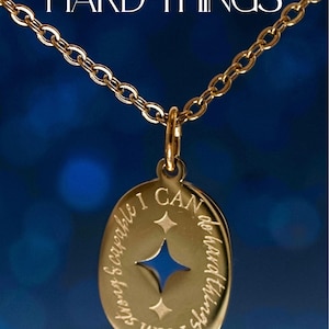 Op de afbeelding: Goudkleurige affirmatieketting met een ketting. De hanger is ovaal van vorm met de woorden "I CAN DO HARD THINGS" en "I AM STRONG & CAPABLE" gegraveerd. Een blauw sterrenpatroon staat in het midden. De woorden "AFFIRMATION NECKLACE" staan onderaan.