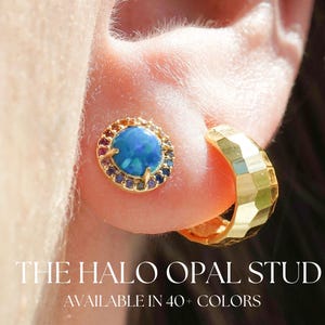 May include: A close-up of a person's ear adorned with a gold hoop earring and a gold stud earring. The stud features a blue opal center surrounded by a halo of colorful gemstones. Text reads: "THE HALO OPAL STUD AVAILABLE IN 40+ COLORS."
