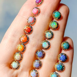 May include: A collection of round stud earrings featuring cabochon opals in various colors including pink, orange, blue, and purple. Each opal is surrounded by a halo of small, multi-colored gemstones set in a gold-toned metal frame.