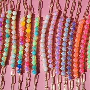 May include: A collection of gold-tone chain bracelets featuring rows of round, iridescent opal-like beads in various colors including pink, blue, orange, purple, and green, arranged in vertical lines against a solid light pink background.