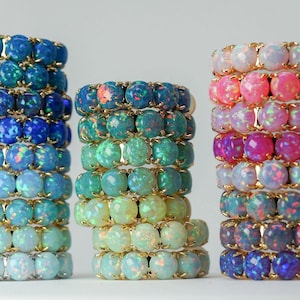 May include: Three vertical stacks of rings featuring round, iridescent cabochon stones set in gold-toned metal. The left stack displays shades of blue, the center stack transitions from blue to green and white, and the right stack shows pink and purple hues.
