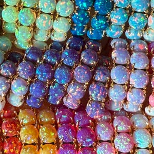 May include: A collection of round, iridescent cabochon stones set in gold-toned metal prongs. The stones display a vibrant spectrum of colors including green, blue, purple, yellow, orange, and pink, each featuring a shimmering, multi-colored play of light.