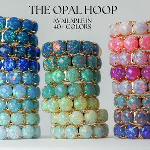 May include: Stacks of opal hoop rings in various colors, including blue, green, and pink. The rings are displayed on a white surface. Text at the top reads "THE OPAL HOOP" and "AVAILABLE IN 40+ COLORS."