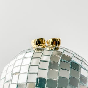 Puede incluir: Dos pendientes de aro dorados con un patrón geométrico descansan sobre una bola de discoteca. Los pendientes tienen un diseño facetado, que refleja la luz. La bola de discoteca está cubierta de pequeñas baldosas de espejo cuadradas.