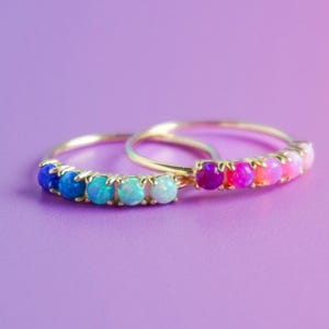 May include: Two gold rings featuring a row of five round cabochon stones each. The first ring displays a gradient of blue and teal stones, while the second ring features a gradient of purple, magenta, and pink stones set in thin gold bands.