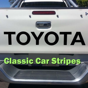 May include: White Toyota pickup truck with black "TOYOTA" lettering on the tailgate. The truck has a black step bumper and a green "Classic Car Stripes" sticker.