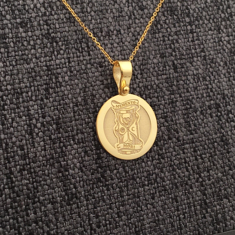 May include: A gold-colored pendant necklace featuring a circular charm with the words "MEMENTO MORI" and an hourglass design with a skull. The necklace has a delicate chain and a simple clasp.