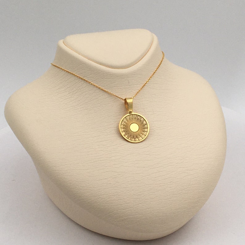 May include: A gold-colored necklace with a sun pendant. The pendant is round with a central circle and radiating lines, resembling a sun. The necklace is displayed on a cream-colored bust, showcasing the jewelry's design and color.