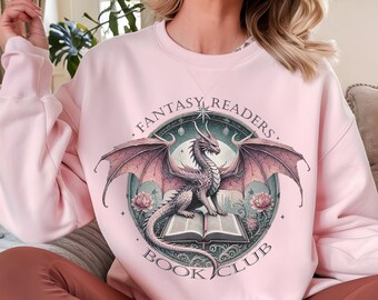 Puede incluir: Una sudadera rosa claro con un gráfico de un dragón posado sobre un libro abierto. El gráfico está rodeado de un círculo con el texto "Fantasy Readers Book Club".