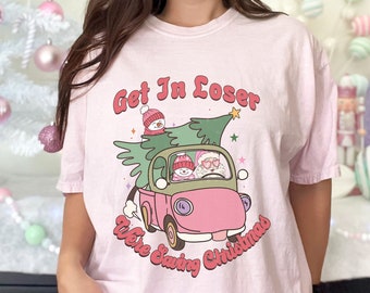 以下が含まれることがあります： ピンクのTシャツに、レトロなスタイルのピンクのトラックがクリスマスツリーを積んでいて、トラックのキャビンには雪だるまが2人とサンタクロースが乗っているというグラフィックが描かれています。Tシャツには「Get In Loser We're Saving Christmas」という文字が書かれています。