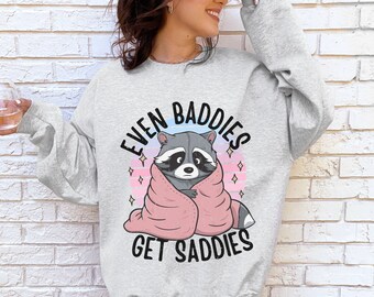 May include: A gray sweatshirt with a cartoon raccoon wearing a pink blanket and the text "Even Baddies Get Saddies".