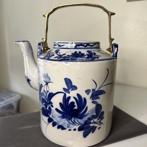 Könnte beinhalten: Eine weiße Teekanne aus Keramik mit einem blauen und weißen Blumendesign und einem Messinggriff.