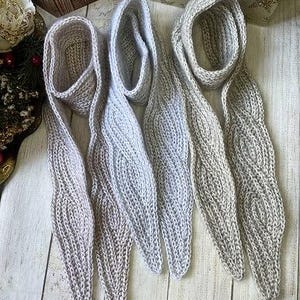 Op de afbeelding: Drie lange, smalle, handgebreide sjaals in verschillende tinten lichtbeige, crème en gebroken wit. Elke sjaal heeft een opvallende, herhalende kabelbreistructuur die over de gehele lengte loopt en eindigt in een taps toelopende punt.