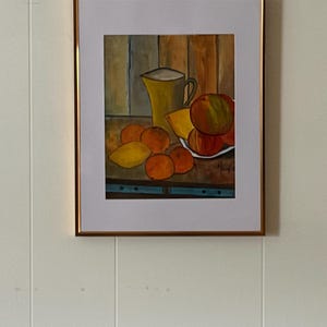 May include: A framed still life painting featuring a yellow pitcher, oranges, lemons, and apples. The artwork has a warm color palette with a wooden background and a gold frame. The painting is mounted on a white wall.