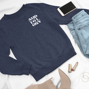 May include: A navy blue sweatshirt with the word "AUDIOLOGY" printed in white on the front. The sweatshirt is laid out on a white surface with a pair of jeans, a cell phone, lipstick, and a pair of earrings.