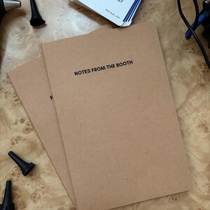 Può includere: Due quaderni di carta marrone con la scritta "NOTES FROM THE BOOTH" sono impilati su una superficie di legno. Sono visibili anche diversi auricolari neri e una guida tascabile per audiologi.