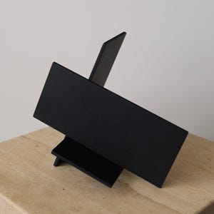May include: A modern, abstract sculpture in matte black. The artwork features intersecting rectangular planes and a minimalist design. The sculpture is displayed on a light-colored, textured surface, creating a contrast with the dark object.