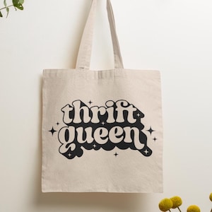 Op de afbeelding: Een witte canvas tote bag met zwarte tekst die "thrift queen" leest in een retro lettertype. De tas hangt aan een houten rek tegen een witte muur.