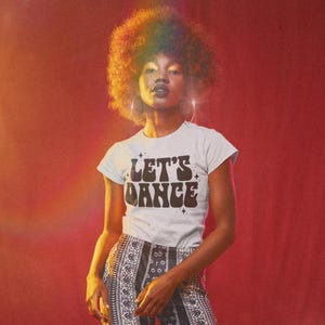 May include: A woman with an afro hairstyle wears a white t-shirt with the text "LET'S DANCE" in a retro font. The shirt has a black and white design with a sparkly effect. She is wearing a patterned skirt and has hoop earrings.
