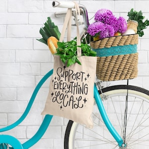 Op de afbeelding: Een turquoise fiets met een rieten mand gevuld met paarse bloemen en een canvas tas met de tekst "Steun alles lokaal".