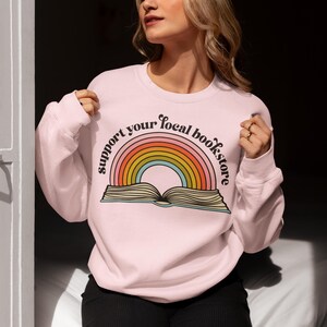 May include: Pink sweatshirt with a rainbow and an open book design. The text "Support your local bookstore" is written in a retro font around the rainbow.