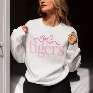 May include: White sweatshirt with the word "tigers" in pink script, topped with a pink bow design. The sweatshirt is a crew neck style and appears to be made of a soft material.