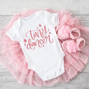 May include: A white baby onesie with the words "tiny dancer" written in pink script. The onesie is on top of a pink tulle tutu and pink sandals with flower embellishments.