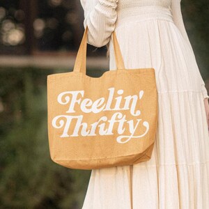 Op de afbeelding: Een bruine tote bag met de tekst "Feelin' Thrifty" in witte letters. De tas is gemaakt van een natuurlijk materiaal en heeft een eenvoudig ontwerp.