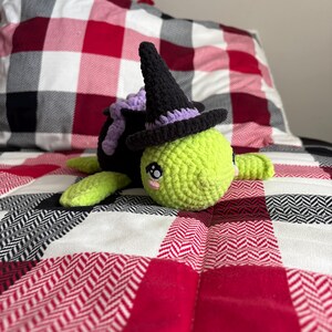 May include: A green crocheted turtle wearing a black witch hat with a purple trim. The turtle is lying on a red and white plaid blanket.