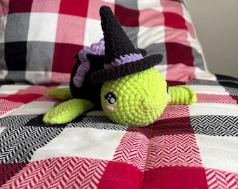 May include: A green crocheted turtle wearing a black witch hat with a purple trim. The turtle is lying on a red and white plaid blanket.