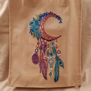 May include: A beige tote bag with a colorful dreamcatcher design featuring a crescent moon, feathers, and beads. The dreamcatcher is made of rhinestones and is a vibrant purple, blue, and green.