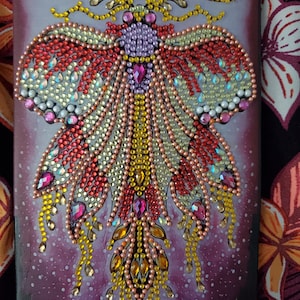 Puede incluir: Un diseño de mariposa morado, rojo y amarillo con acentos iridiscentes sobre un fondo morado oscuro. El diseño está hecho de pequeñas cuentas de colores.