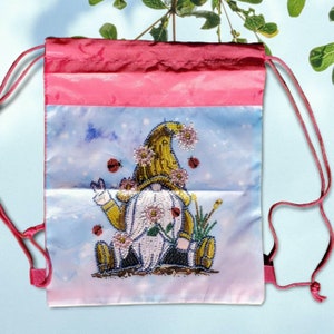 以下が含まれることがあります： 黄色い帽子と花をつけた漫画のノームが描かれた、白と青のデザインのピンクの巾着袋。ノームは花とテントウムシに囲まれています。