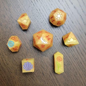 May include: Set of seven gold-colored polyhedral dice with red numbering and themed icons. The icons include a seashell, a rose, a crown, a sun, a magic lamp, a spiral, and a glass slipper. The dice are for tabletop gaming.