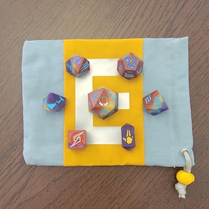 Puede incluir: Un conjunto de siete dados coloridos con varios símbolos y números en cada cara. Los dados están en una bolsa de cordón azul y amarillo.
