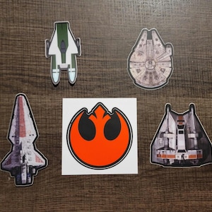 May include: A collection of Star Wars-themed stickers featuring various spacecraft and the Rebel Alliance symbol. The stickers include the Millennium Falcon, a green and white ship, and a red and white ship. The Rebel Alliance symbol is orange and black.