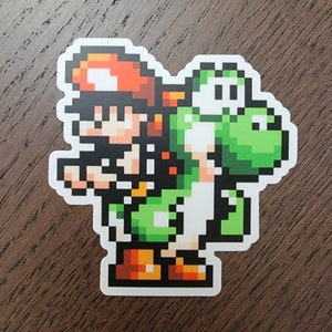 Può includere: Un adesivo pixelato con un Yoshi verde che trasporta un personaggio con un berretto rosso e capelli castani. L'adesivo è su una superficie di legno marrone.