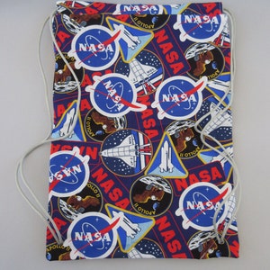 May include: A blue drawstring bag with a repeating pattern of NASA logos, space shuttle illustrations, and the word "NASA" in red. The bag has a white drawstring closure.