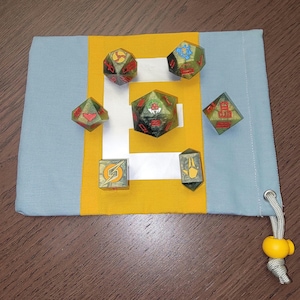 May include: A set of seven green and yellow dice with red and white symbols on each face. The dice are in a grey and yellow drawstring bag.