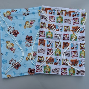 May include: Two drawstring bags with Animal Crossing characters. The bag on the left is light blue with a white drawstring and features Tom Nook, Isabelle, and other characters. The bag on the right is white with a white drawstring and features a repeating pattern of Animal Crossing characters in colorful squares.