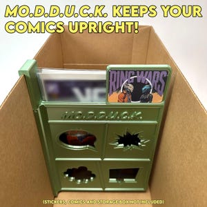 May include: A green plastic comic book storage organizer with the text "MO.D.D.U.C.K. KEEPS YOUR COMICS UPRIGHT!" and "MO.D.D.U.G.K." on the front. The organizer has a comic book inside and a sticker on the top that says "RING WARS".